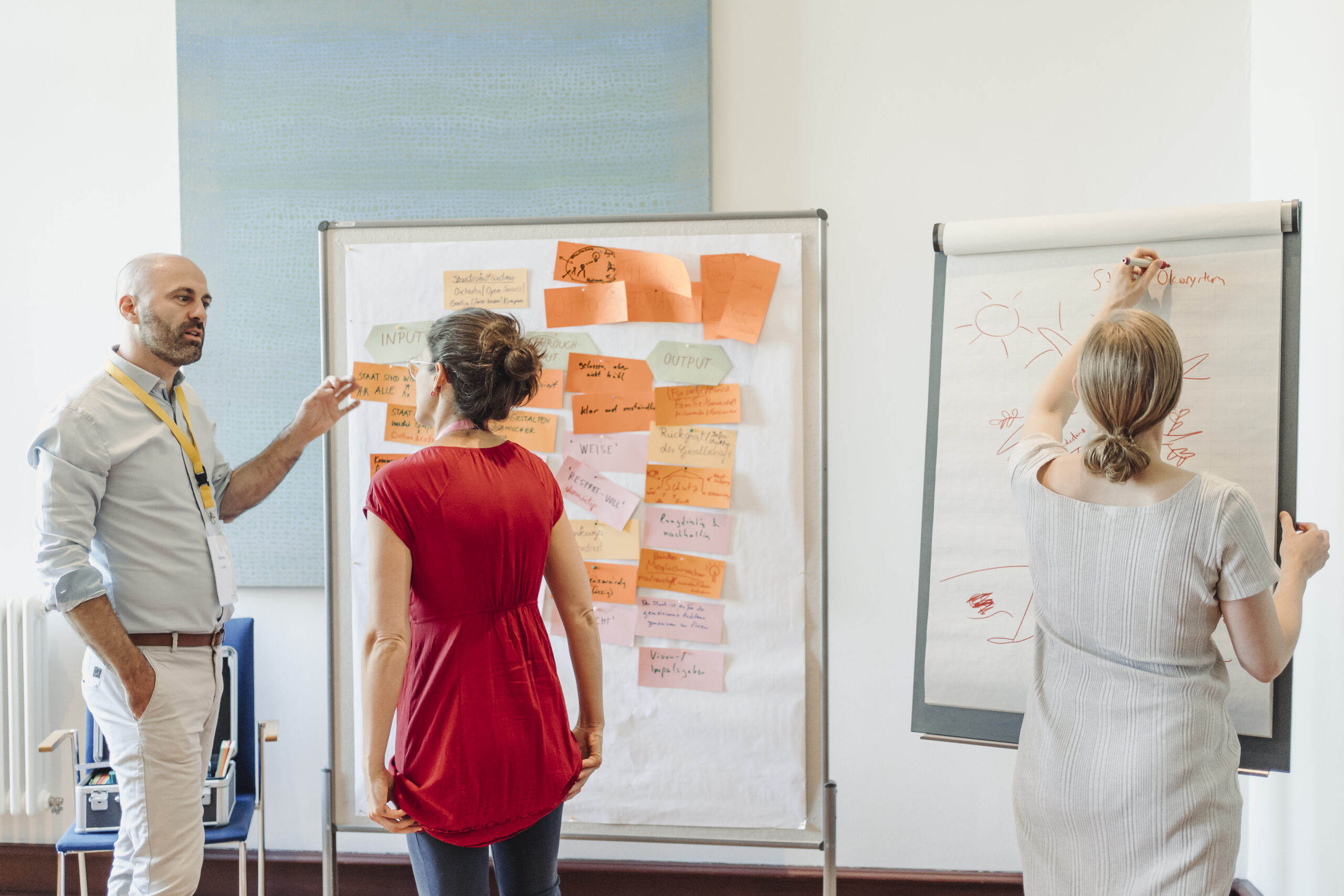 Eine Frau und ein Mann stehen vor einer Pinnwand, Judith schreibt auf eine Präsentationstafel Notizen